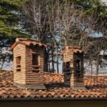 Two rustic brick chimneys on a red-tiled roof with a winter forest backdrop.